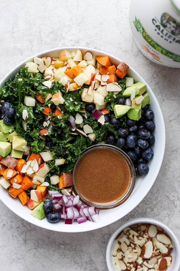 Bowl of massage kale salad that is filled with sweet potatoes, blueberries, avocado, apple and slivered almonds. 