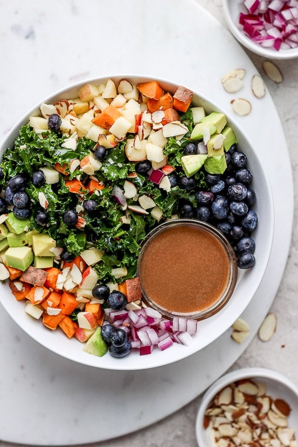 Bowl of massage kale salad that is filled with sweet potatoes, blueberries, avocado, apple and slivered almonds. 