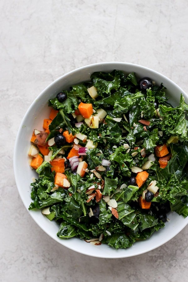 Bowl of massage kale salad that is filled with sweet potatoes, blueberries, avocado, apple and slivered almonds. 