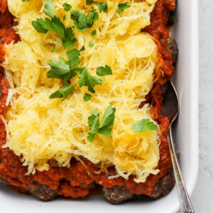 A white baking dish of baked spaghetti squash and meatballs with a spoon sticking out the side.