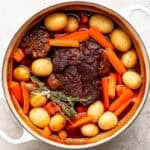 A top down shot of a beef pot roast in a Dutch oven surrounded by fresh herbs, baby potatoes and carrots.