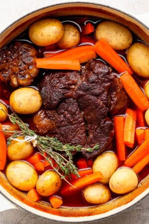 A top down shot of a beef pot roast in a Dutch oven surrounded by fresh herbs, baby potatoes and carrots.