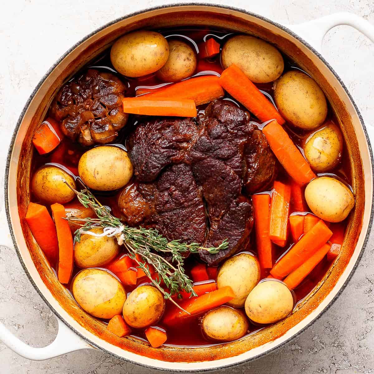 A top down shot of a beef pot roast in a Dutch oven surrounded by fresh herbs, baby potatoes and carrots.