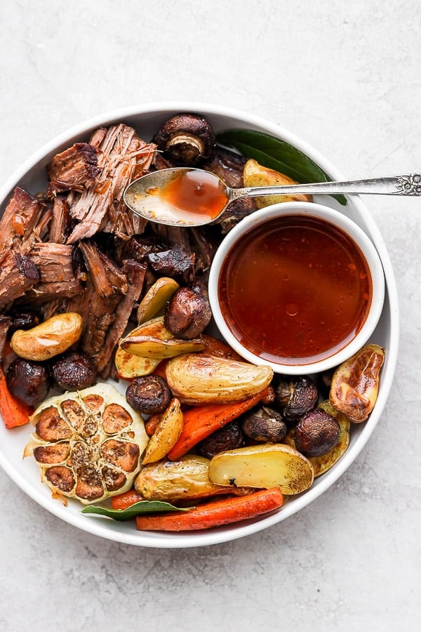 Some of the pot roast and roasted vegetables on a white plate with a small dish of au jus.