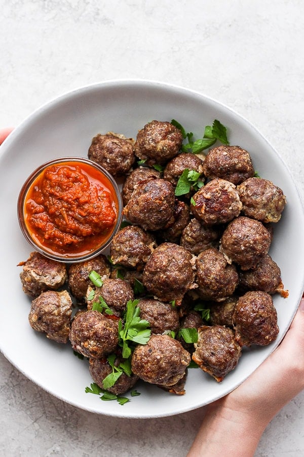 Oven baked meatballs in a large white bowl with a small dish of marinara sauce.