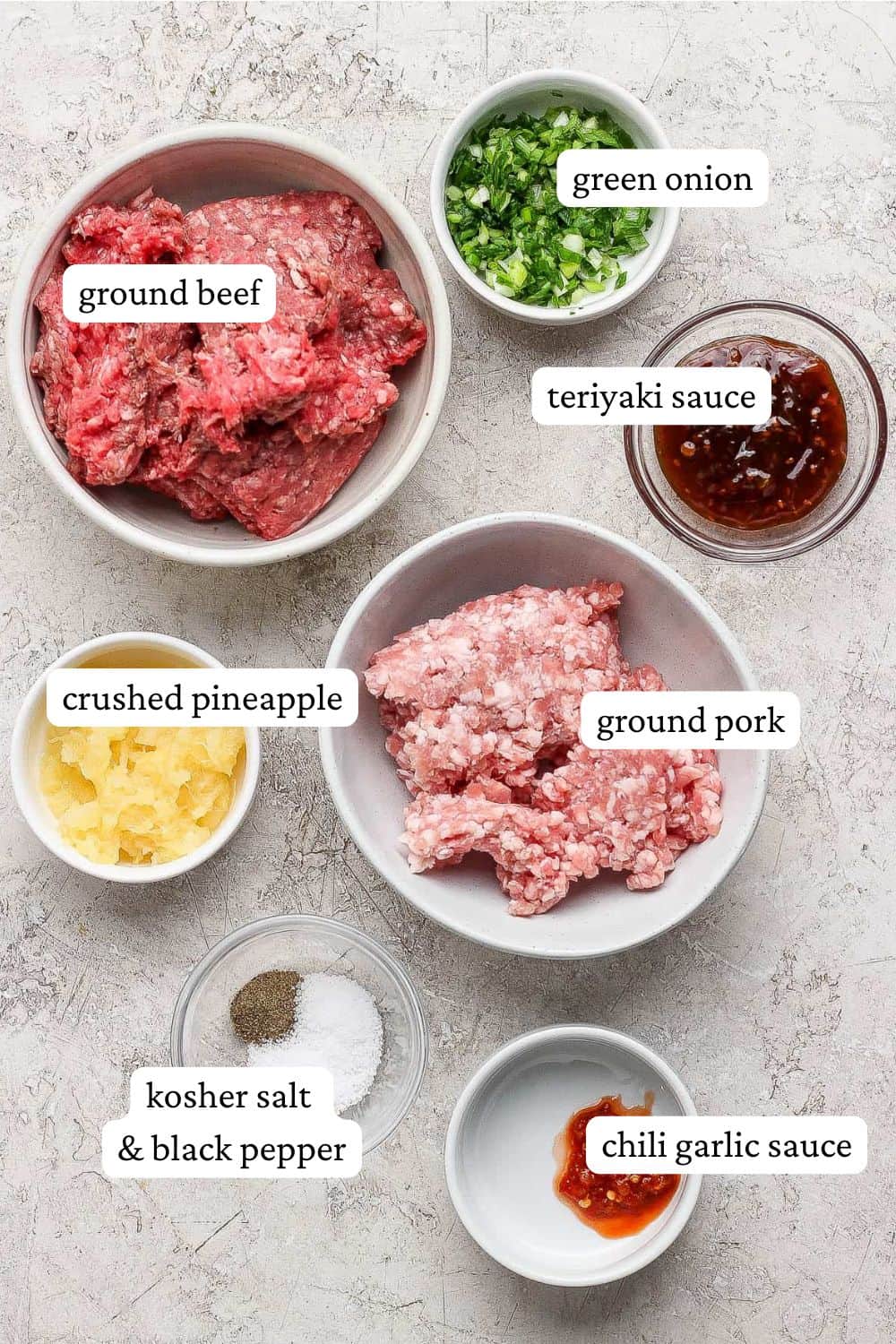 Pineapple teriyaki meatball ingredients on a counter in individual bowls; ground beef, ground pork, green onions, teriyaki sauce, crushed pineapple, kosher salt, ground black pepper, and chili garlic sauce.