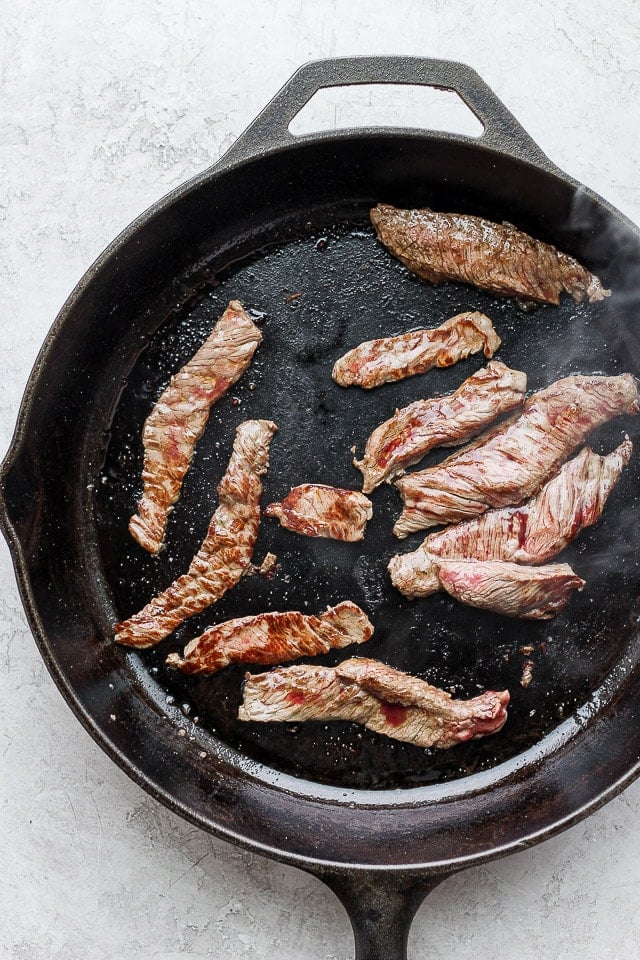 Steak strips sautéing in a large cast iron skillet.