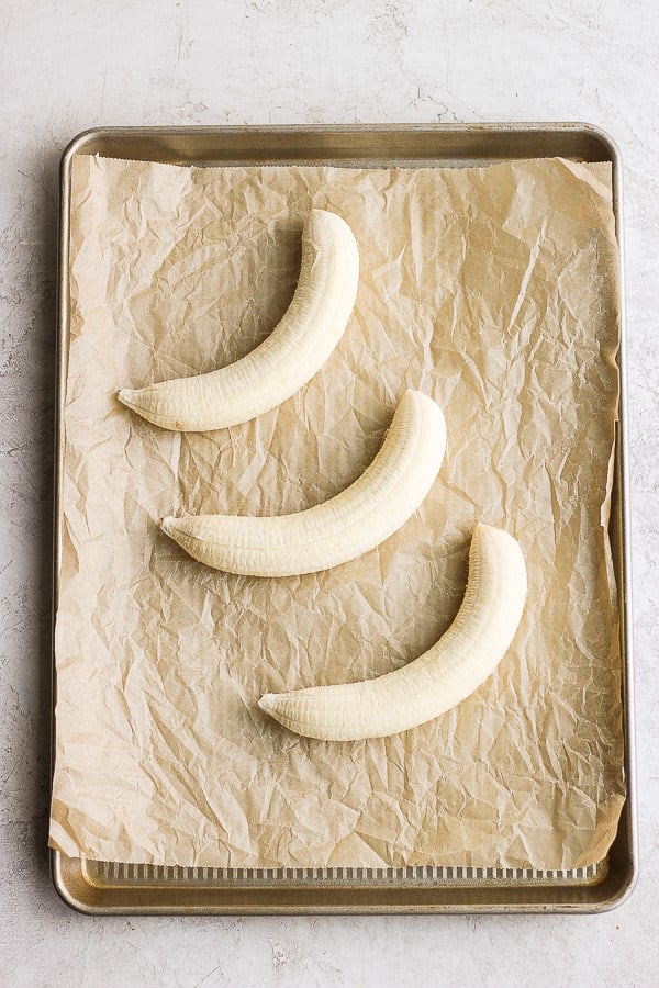 Three peeled bananas on a parchment-lined baking sheet.
