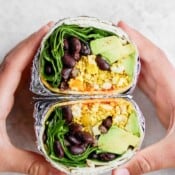 Two hands holding one breakfast burrito that has been cut in half.