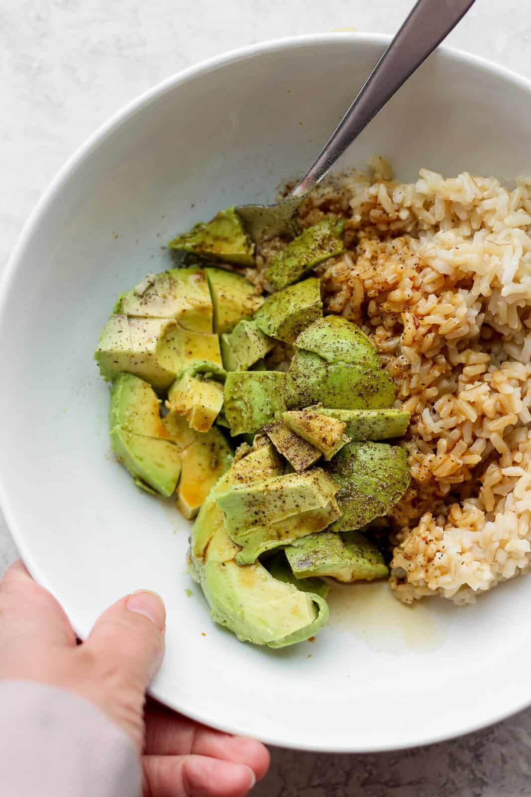 A hand holding a bowl of avocado, rice and soy sauce.