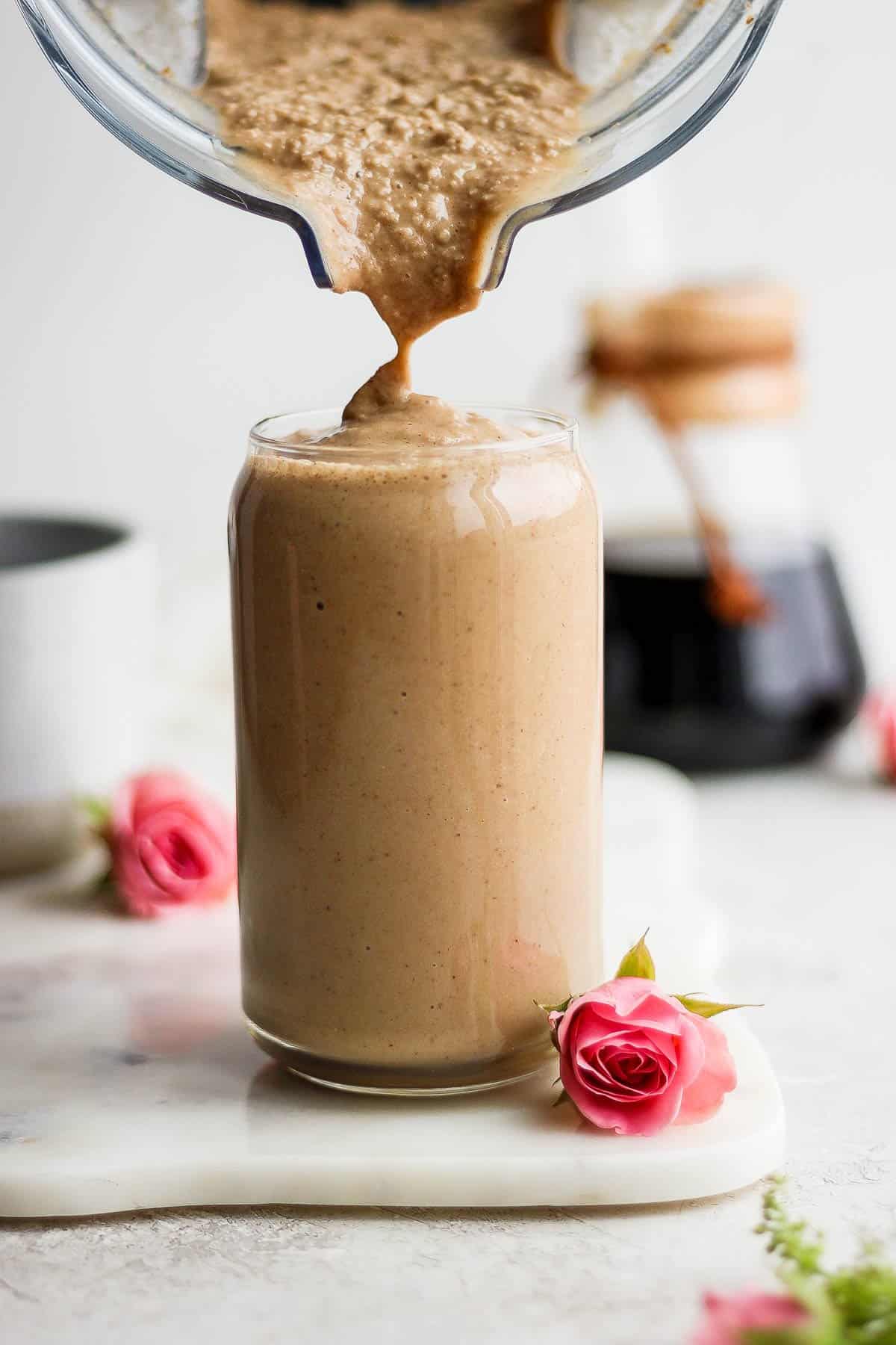 Someone pouring the smoothie into a glass which is sitting on a marble board next to pink flowers.