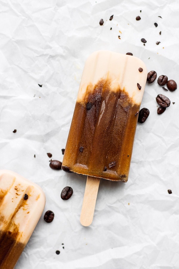 Two coffee popsicles on a piece of crumpled parchment paper with coffee beans around. 