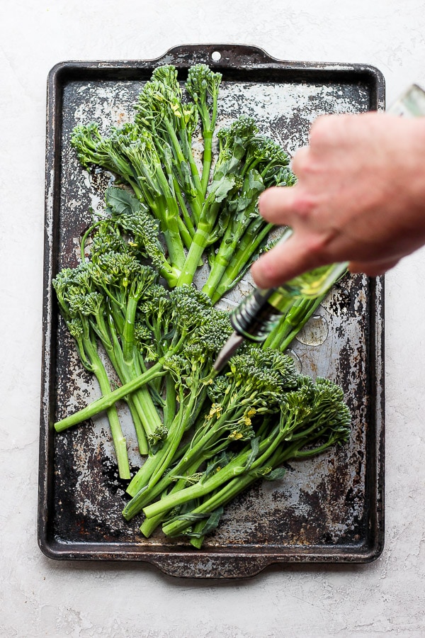 Raw broccolini on a cookie sheet and someone drizzling olive oil on top. 