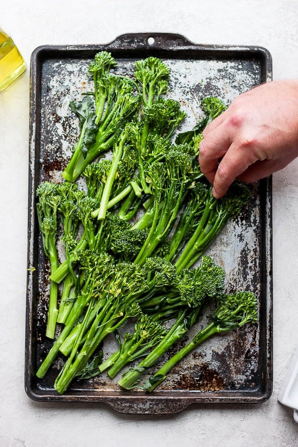 Raw broccolini on a cookie sheet and hands rubbing in the olive oil.