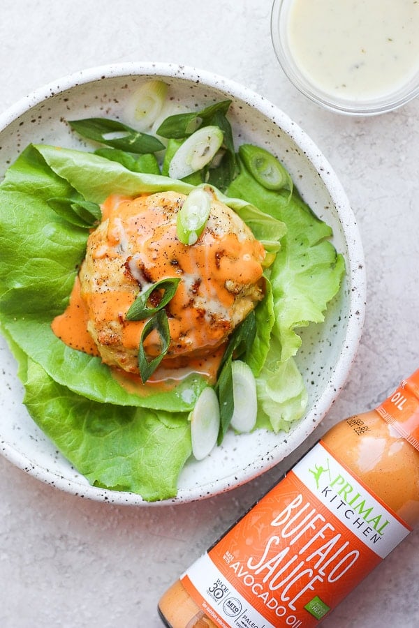 A buffalo chicken burger sitting in a bed of butter lettuce in a shallow bowl with a bottle of buffalo sauce next to it. 