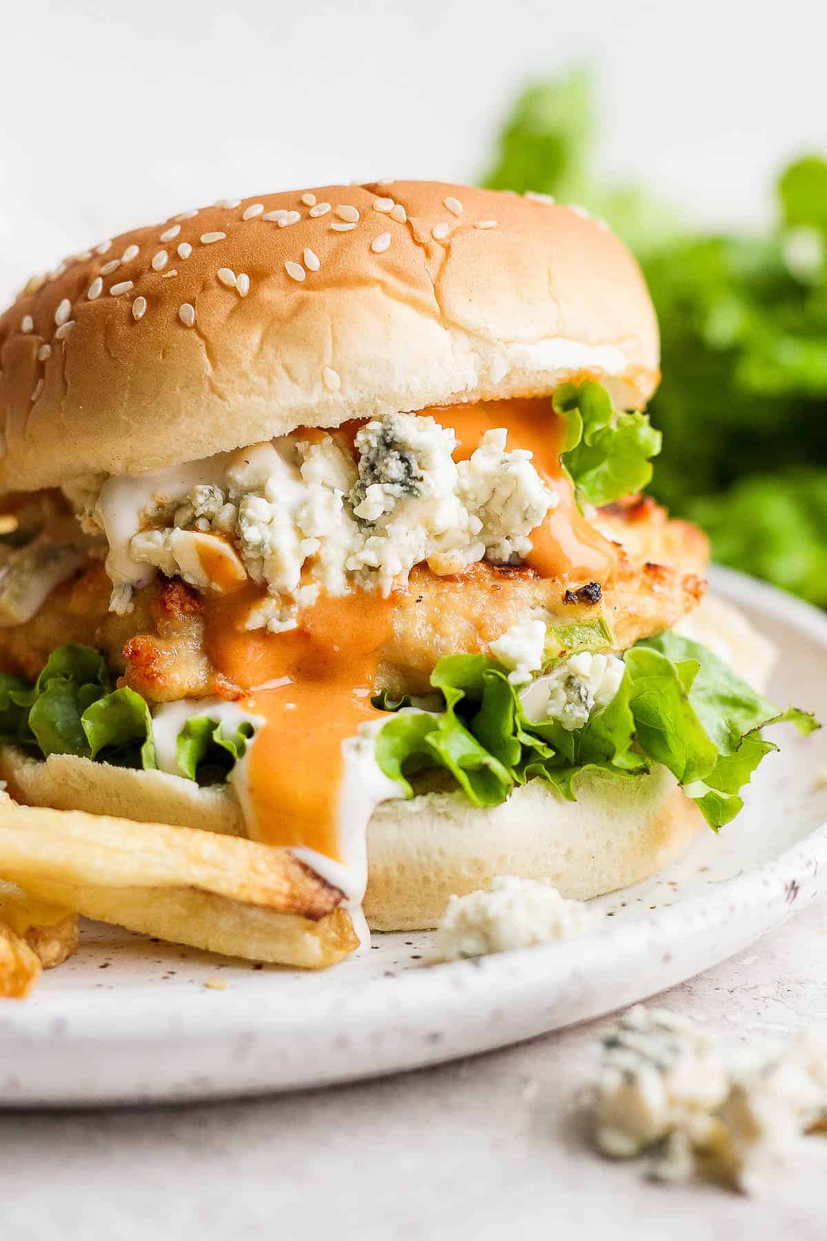 A buffalo chicken burger on a bun on a plate with french fries. 