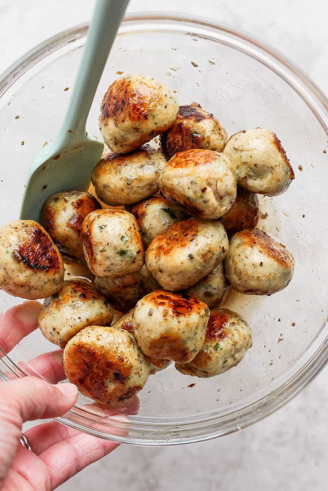 Someone holding a glass bowl with cooked Greek chicken meatballs tossed in Greek vinaigrette inside and a spatula sticking out. 