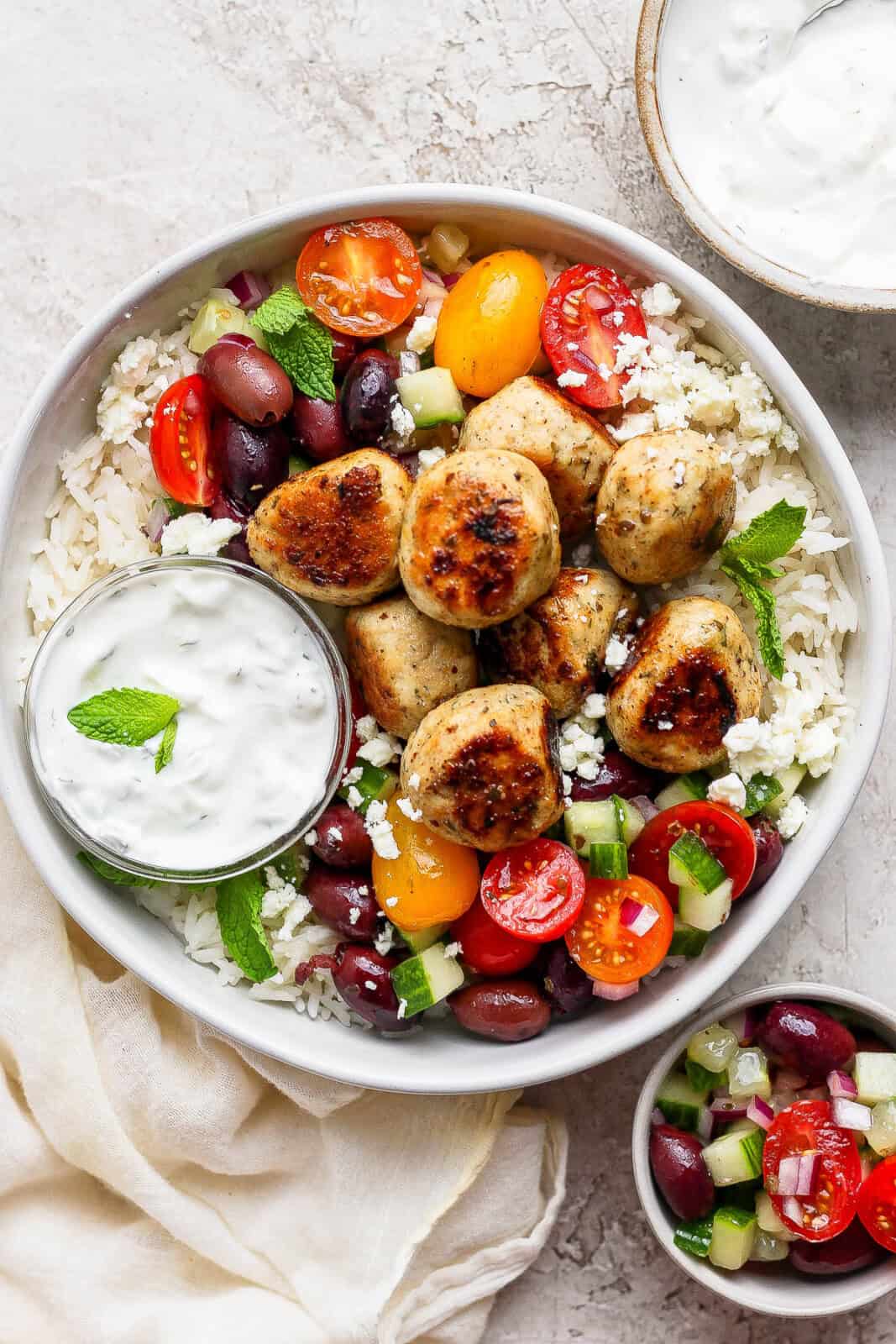 A prepared bowl of cooked Greek chicken meatballs, rice, feta, Greek salad and tzatziki sauce. 