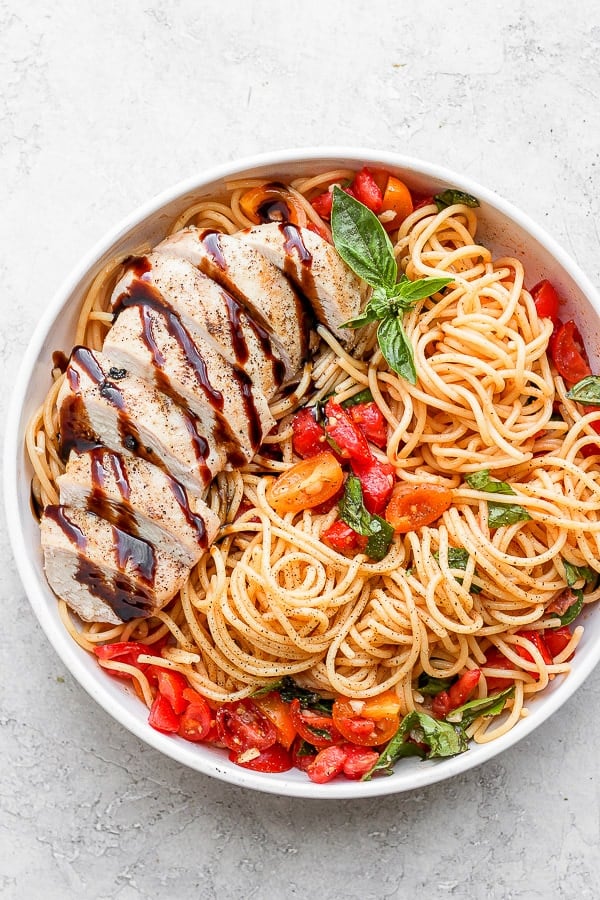 A bowl of bruschetta chicken pasta with tomatoes, fresh basil and a grilled chicken breast drizzled with balsamic glaze.
