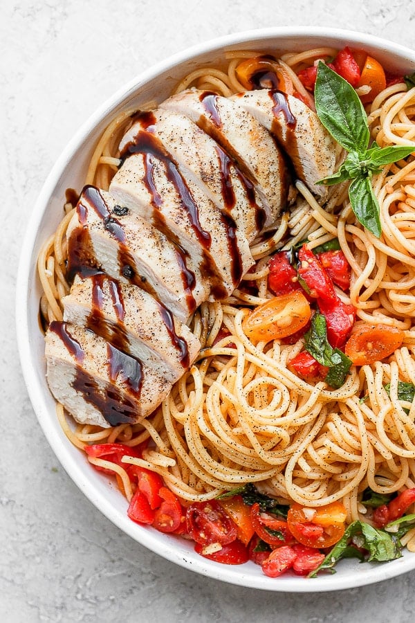A bowl of bruschetta chicken pasta with tomatoes, fresh basil and a grilled chicken breast drizzled with balsamic glaze.