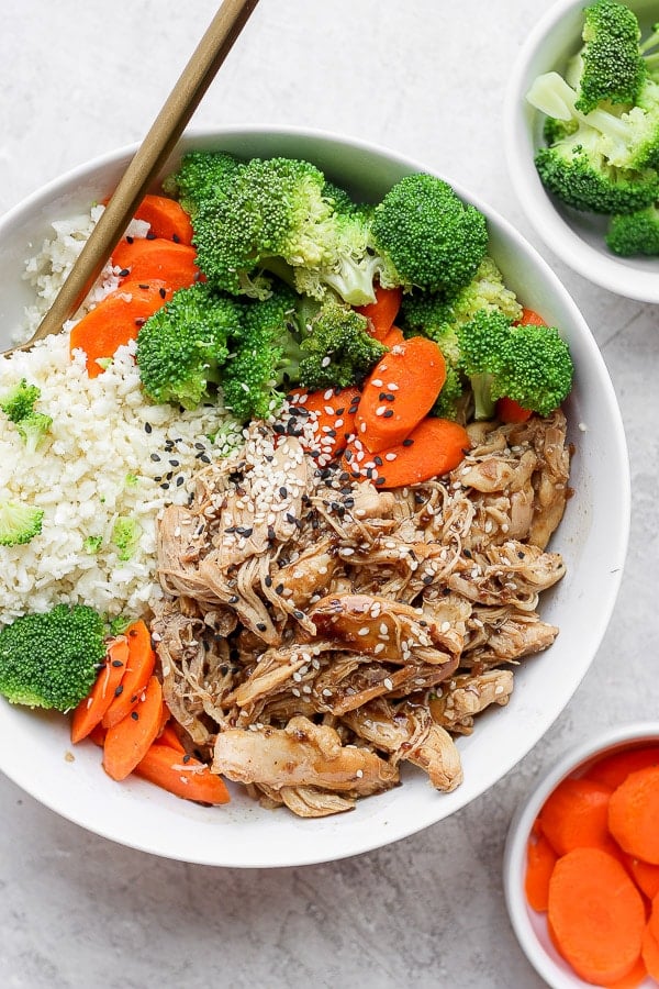 A teriyaki chicken bowl in a white bowl and small bowls of vegetables on the side.