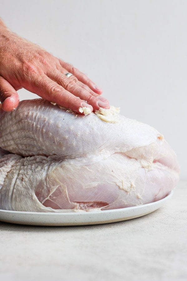 A hand rubbing softened butter all over a raw turkey breast. 