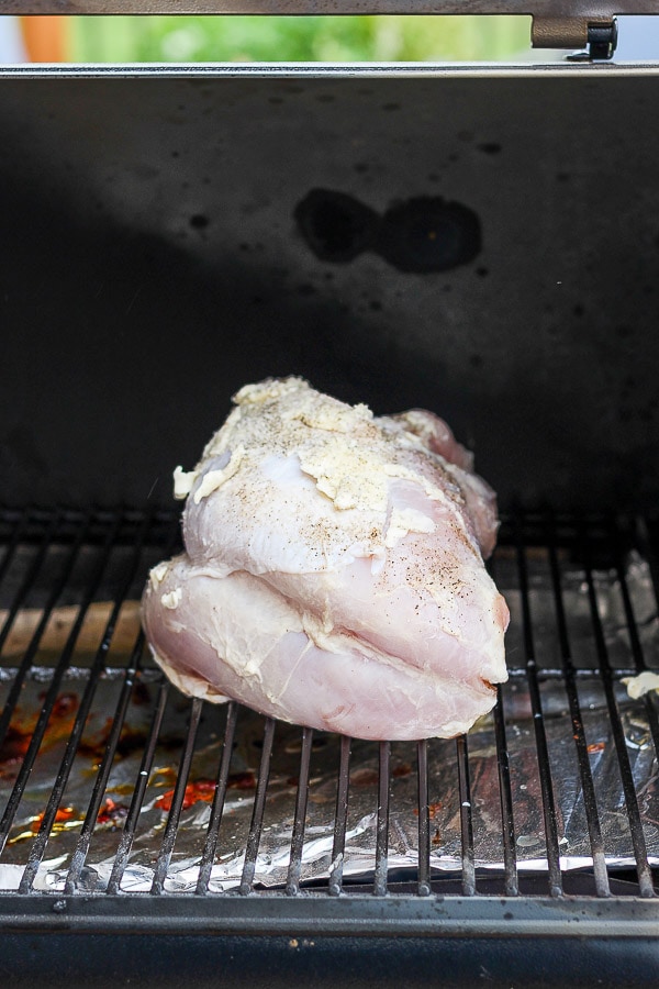 A raw turkey breast covered in soft butter and seasoned with salt and pepper sitting on the grates of an open smoker. 