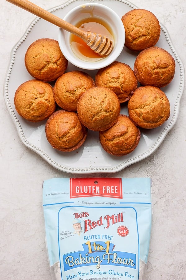 A pile of cornbread muffins on a white plate with a small bowl of honey next to a bag of Bob's Red Mill 1 to 1 baking flour.