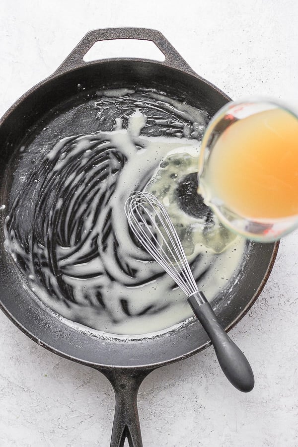 Cast iron skillet with a whisk combining melted ghee and tapioca starch and chicken broth being poured into the pan. 