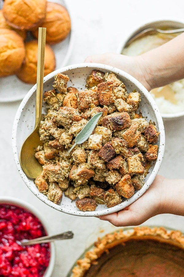 Two hands holding a bowl of gluten free stuffing over a table full of thanksgiving food. 
