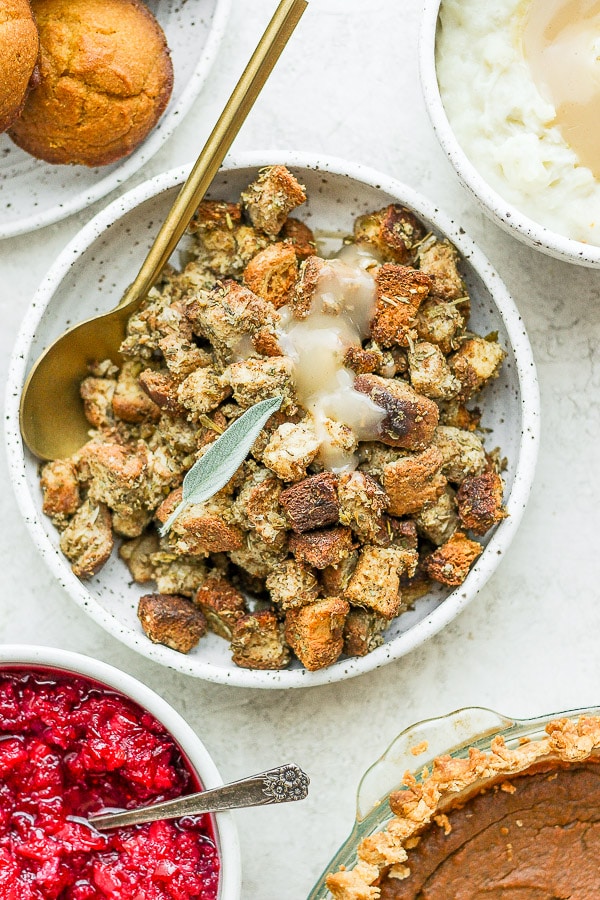 A bowl of gluten free stuffing with gravy poured on top and surrounded by Thanksgiving food. 