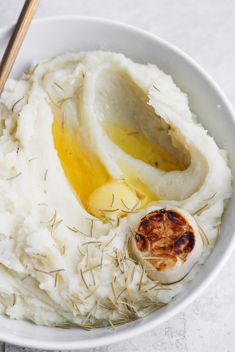 A bowl of mashed potatoes with melted ghee, a head of roasted garlic, rosemary, and a spoon.