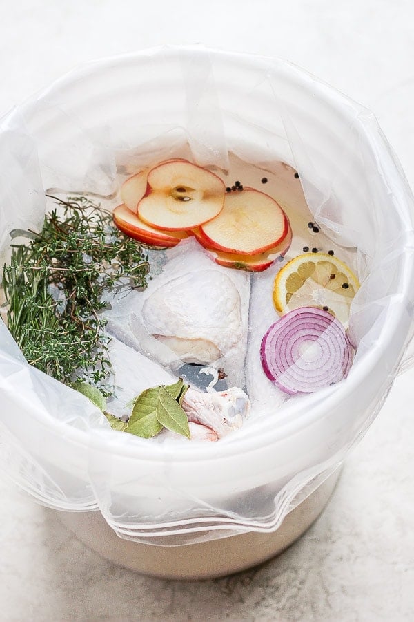 A large bucket with a brine bag in it with a raw turkey sitting in a brine with herbs, apples, onion and lemon. 