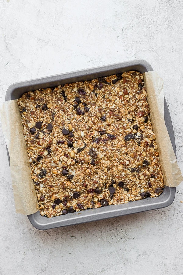 A 8x8 pan with a piece of parchment in it and the trail mix bar ingredients pressed down. 
