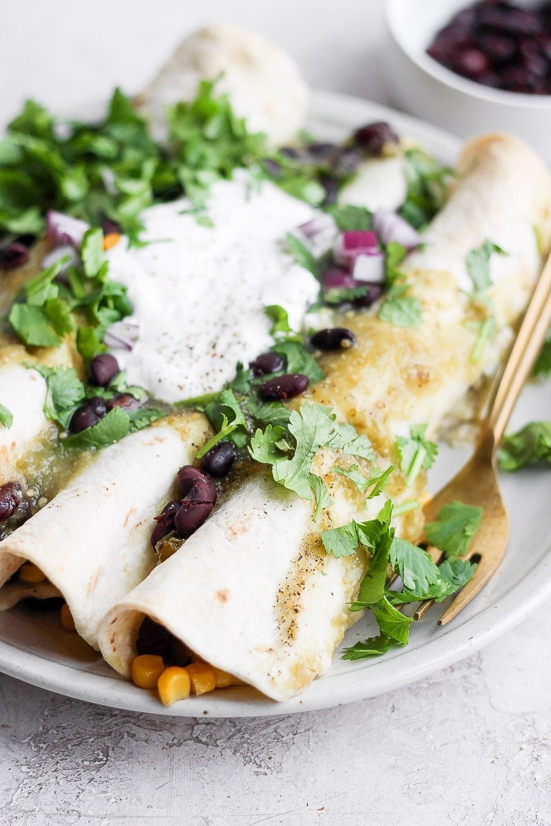 Side angle of a plate with three vegan enchiladas topped with fresh cilantro, dairy free sour cream and red onions. 