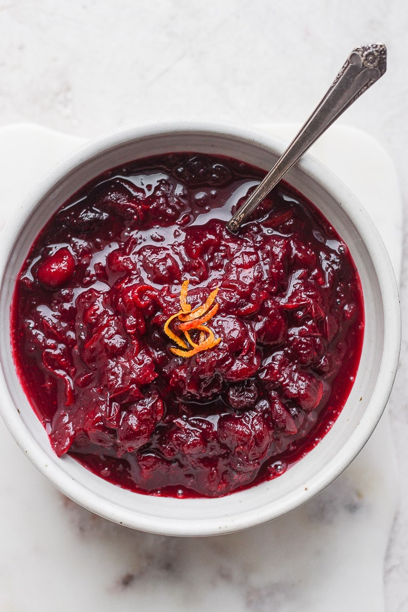Cranberry orange sauce in a bowl with orange zest on top and a spoon sticking out. 