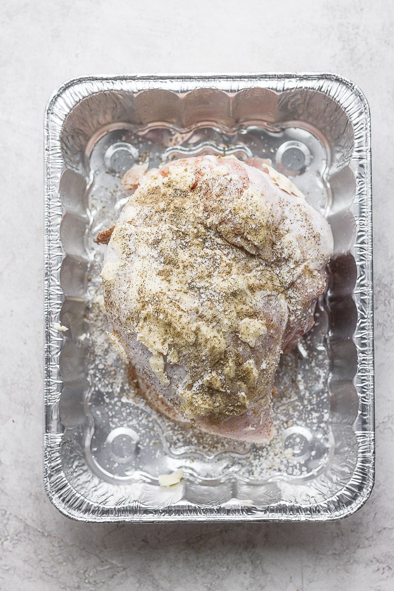 Turkey breast in an aluminum tray that has been covered in butter, salt, and pepper.