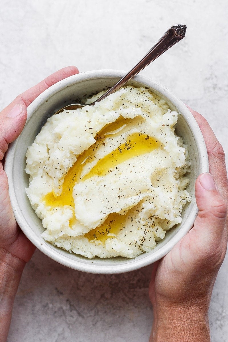 Hands holding a bowl of mashed potatoes with melted ghee, salt, pepper, and a spoon.