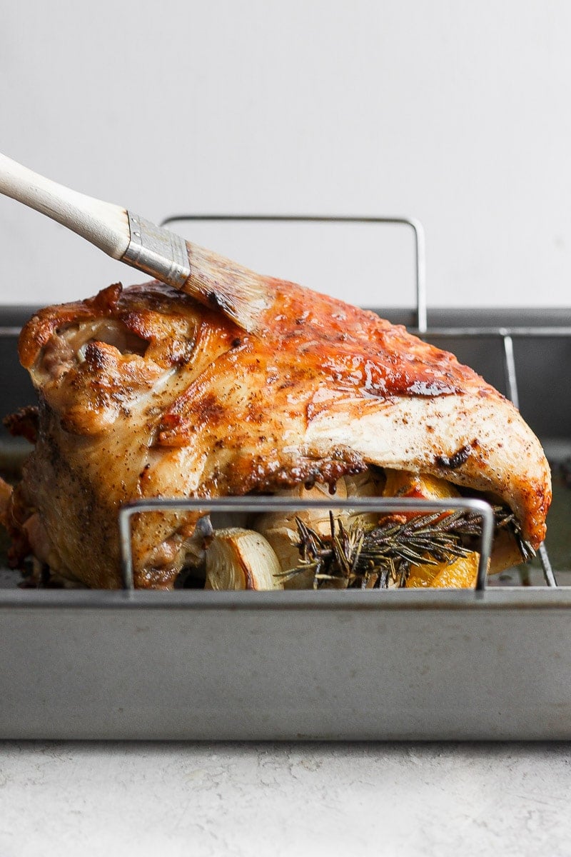 Side view of a cooked, brown turkey breast in a roasting pan and someone basting it with a brush. 