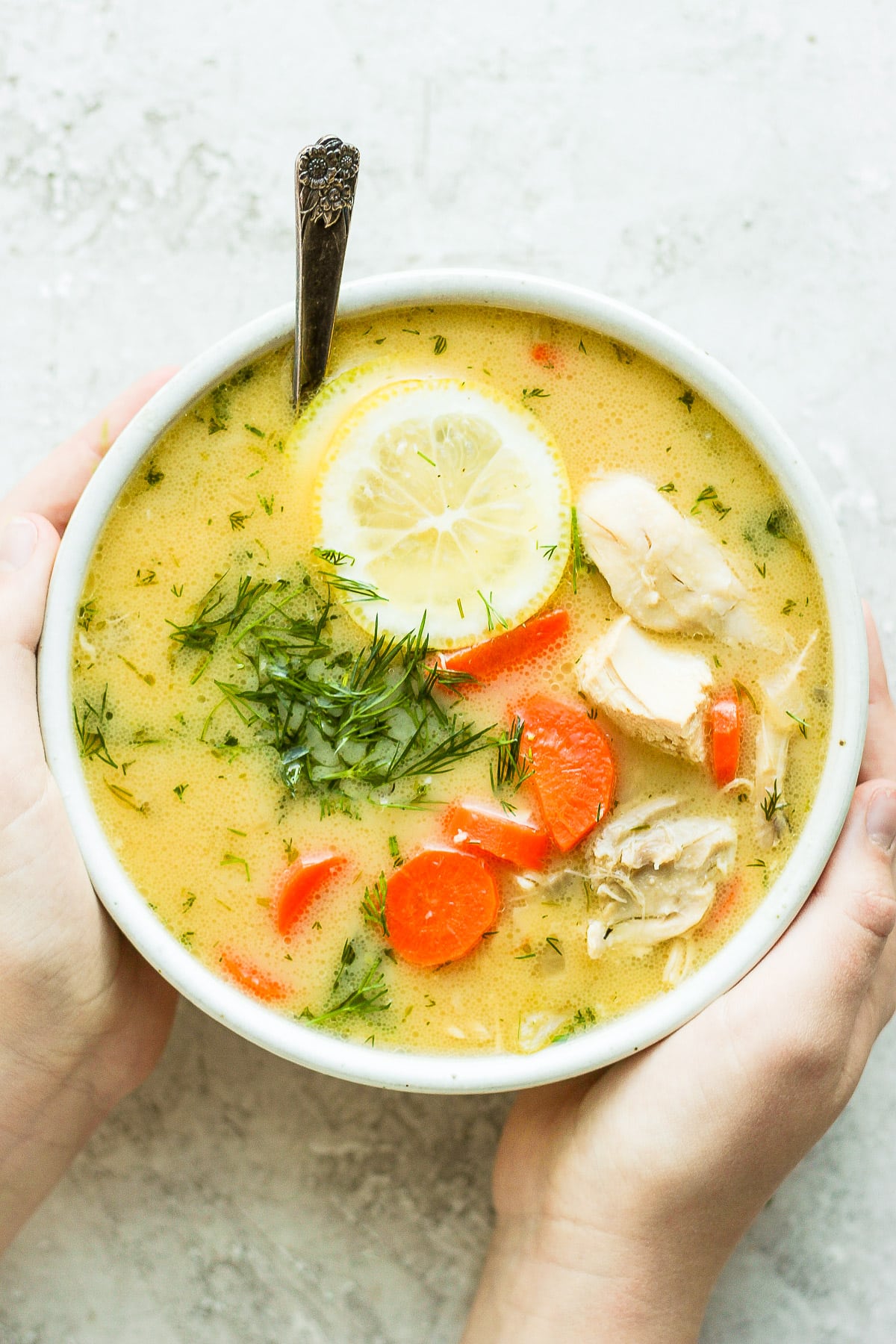 Two hands holding a bowl of greek lemon chicken soup.