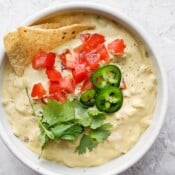 A white bowl full of vegan queso with two tortilla chips sticking out, diced tomatoes, jalapeño slices, and fresh cilantro.