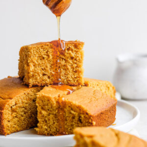 Squares of gluten-free cornbread piled on a white plate with honey being drizzled on top.