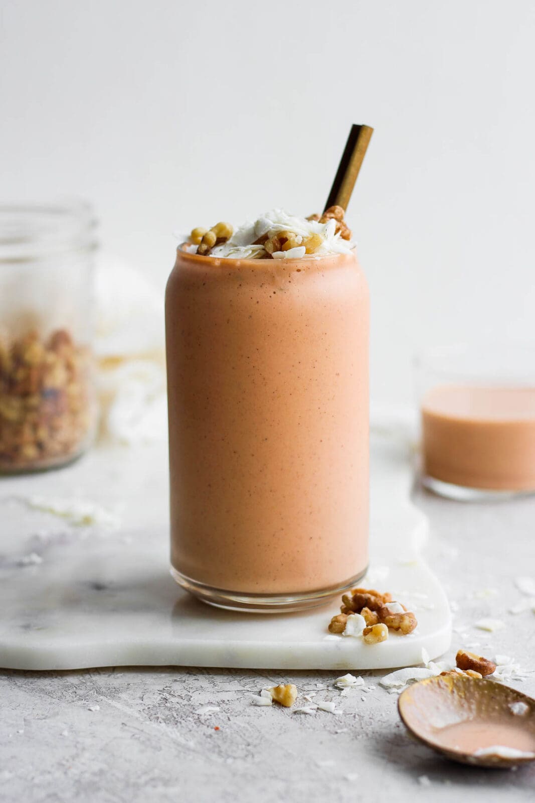 Carrot cake smoothie in a glass topped with coconut and walnuts with a gold spoon sticking out. 