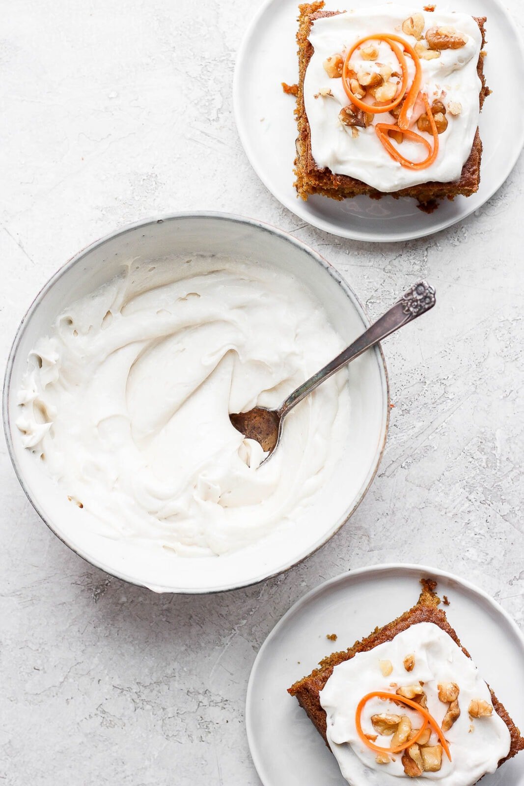 Bowl of dairy free cream cheese frosting with spoon and 2 plates of carrot cake with frosting.