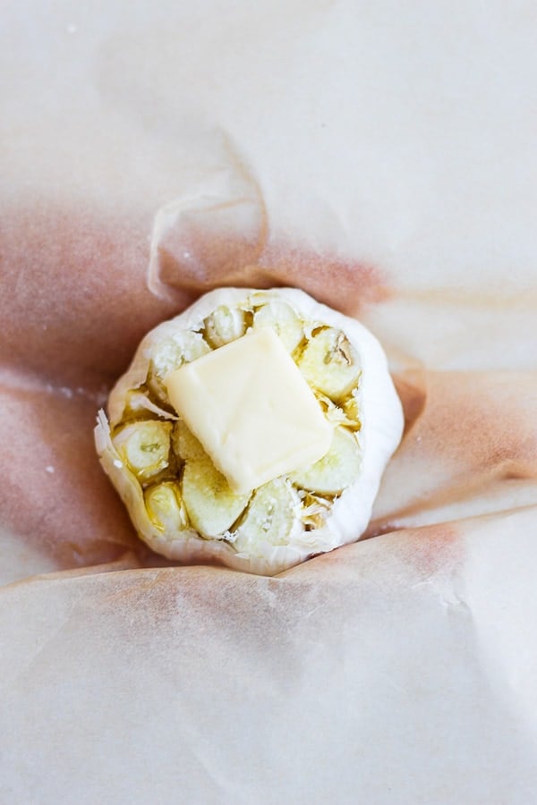 A head of garlic with olive oil, salt, and butter on a piece of parchment paper.