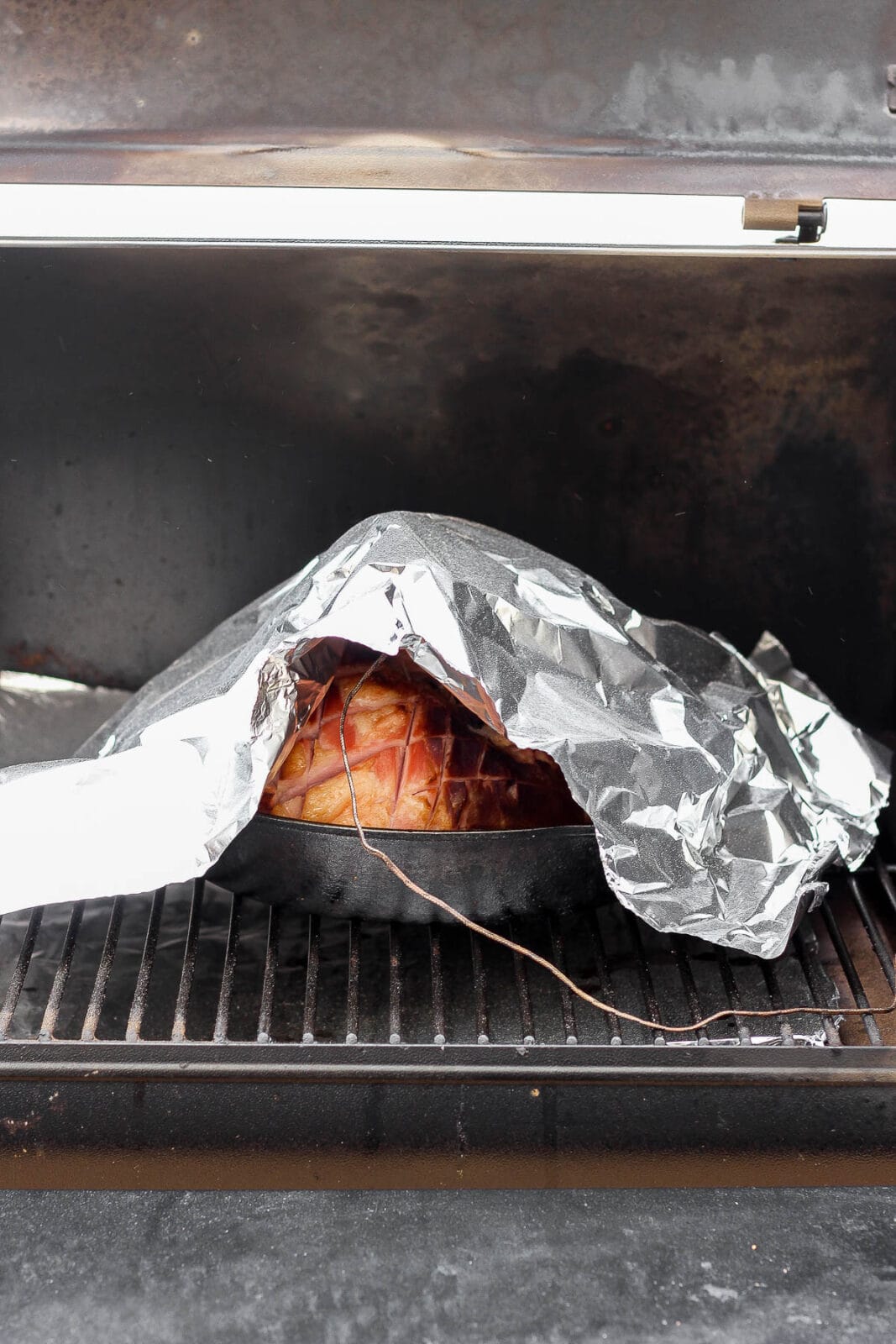 Ham in a cast iron skillet in a smoker with foil on the top and a meat thermometer inserted in the ham.