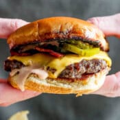 Someone holding a smash burger with bacon, pickles, cheese and burger sauce on a plate with chips.