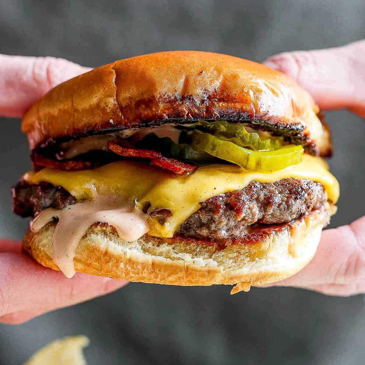 Someone holding a smash burger with bacon, pickles, cheese and burger sauce on a plate with chips.