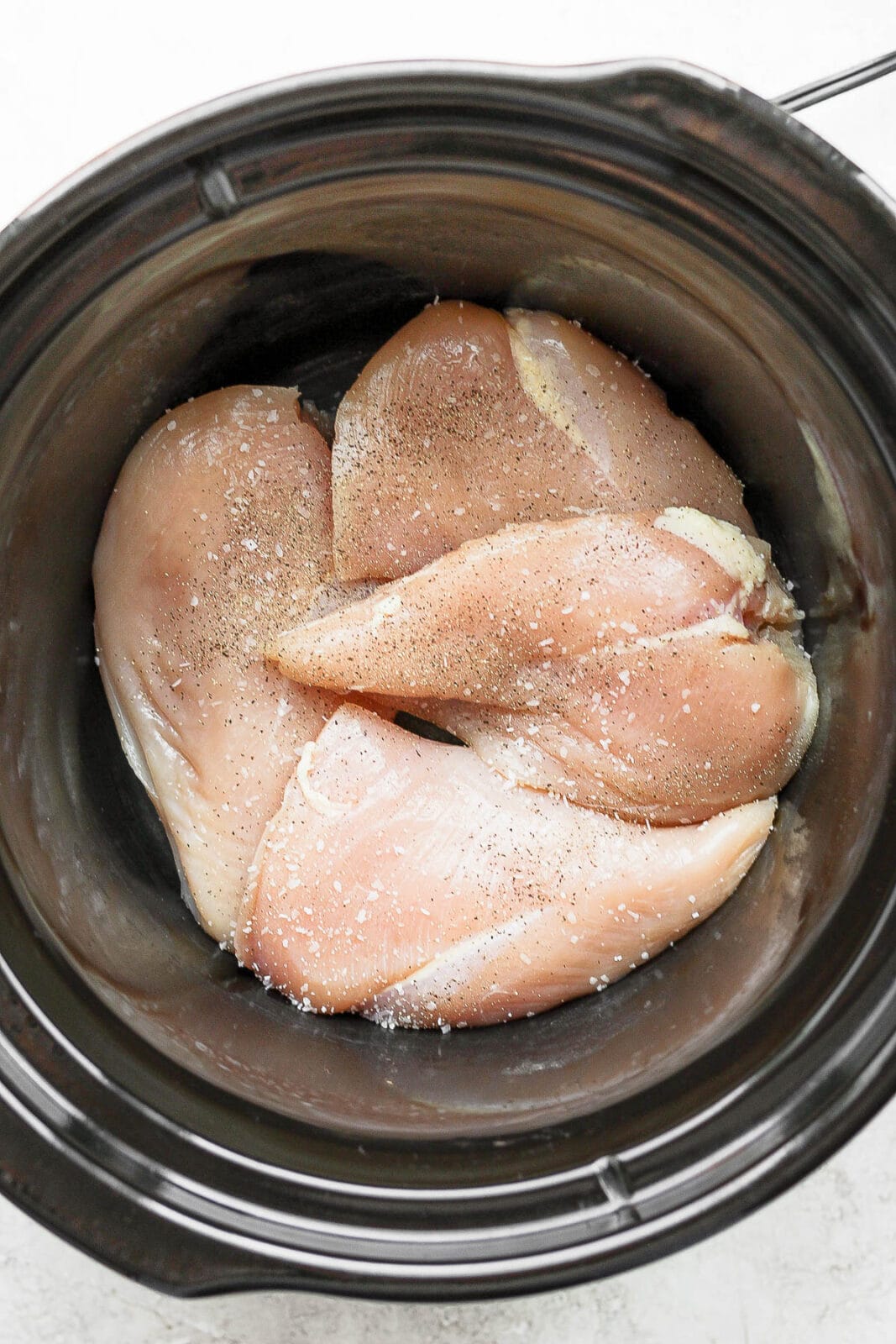 Slow cooker with raw chicken breasts, salt, and pepper.