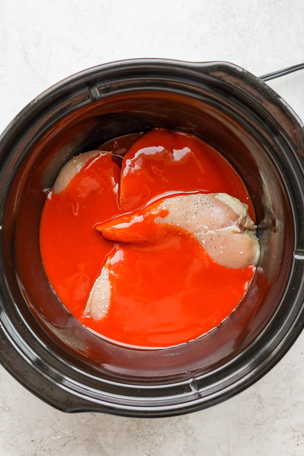 Raw chicken breasts in a slow cooker with buffalo sauce.